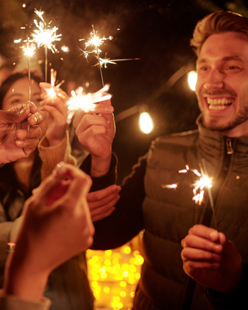happy young intercultural friends in birthday caps holding bengal lights having fun and enjoying outdoor party in the evening